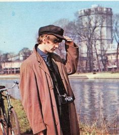 a man standing next to a river wearing a coat and tie with a bike parked in front of him