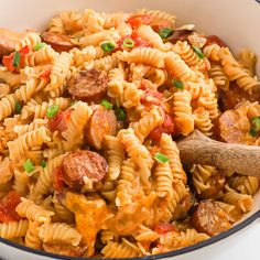 a pasta dish with sausage, tomato sauce and green onions in a white bowl next to a wooden spoon