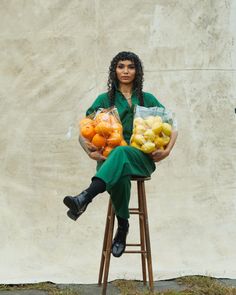 a woman is sitting on a stool holding bags of oranges and apples in front of a wall