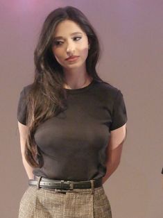 a woman standing in front of a white wall wearing a black shirt and tan pants