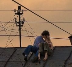 two people sitting on the roof of a building talking on their cell phones at sunset