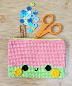 a crocheted pink and green pencil case with buttons on the side, sitting on a wooden surface