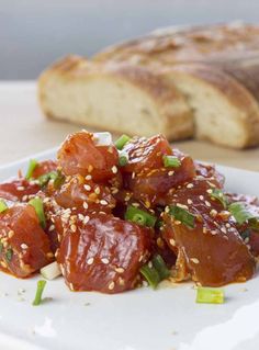 a white plate topped with meat covered in sauce and sesame seeds next to a loaf of bread