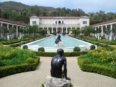 a statue sitting in front of a fountain surrounded by bushes and flowers with a building in the background