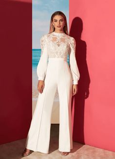 a woman standing in front of a pink wall wearing white pants and a lace top