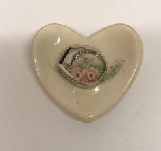 a small white heart shaped object with flowers on it's side and a ring in the middle