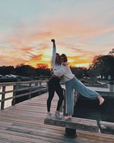 two women standing on a dock with their arms around each other as the sun sets