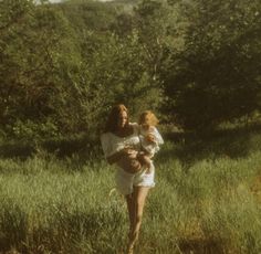 a woman holding a baby in her arms while walking through tall grass with trees behind her