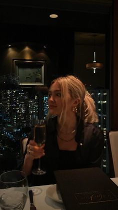 a woman sitting at a table with a glass of wine in front of her and the city lights behind her