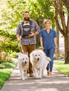 a man and woman walking two white dogs down a sidewalk with trees in the background