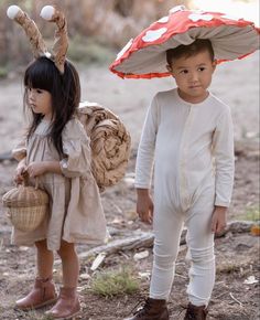 two young children standing under an umbrella in the dirt, one holding a basket and the other wearing bunny ears