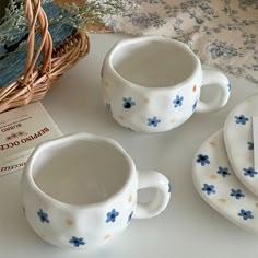 three white cups and saucers with blue flowers on them, next to a wicker basket