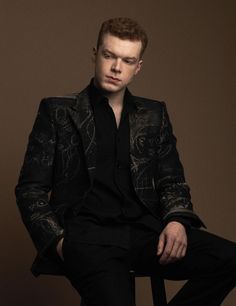 a man sitting in a chair wearing a black shirt and blazer with his hands on his knees