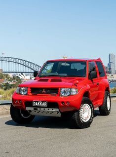 a bright red suv parked in front of a bridge with sydney harbour in the background