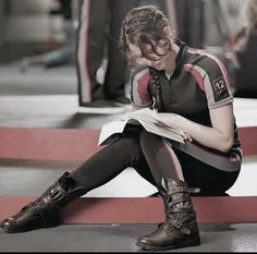 a young woman sitting on the ground reading a book with her legs crossed and wearing boots