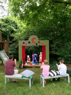 children are sitting on benches in front of an outdoor stage with clowns and stars