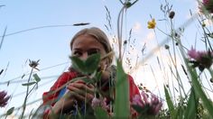 a woman standing in the middle of tall grass with flowers around her and looking up into the sky