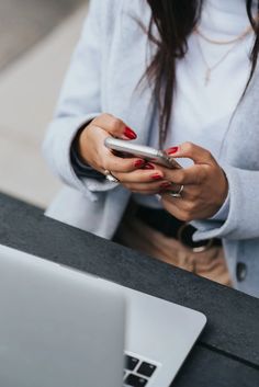 a woman using her cell phone while sitting at a table in front of a laptop