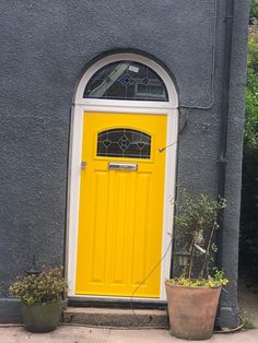 a yellow door sits in front of a gray building with potted plants on the sidewalk