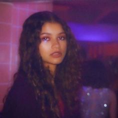 a woman with long curly hair standing in front of a pink wall and looking at the camera