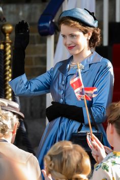 a woman in a blue dress and hat waves to the crowd as she stands with other people