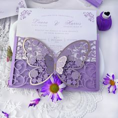 a purple and gold butterfly wedding card on a lace doily with flowers next to it