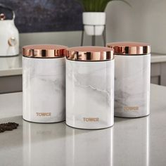 three white and rose gold canisters sitting on a counter top next to a potted plant