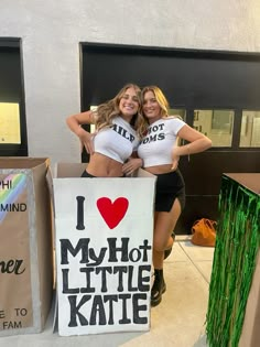 two women in white shirts and black shorts standing next to a sign that says i love my hot little kate