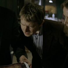 two men looking at an open book in a dark room with other people around them