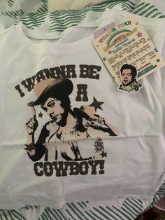 a white t - shirt with the words i wanna be it cowboy on it and a sticker next to it