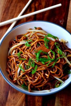 a white bowl filled with noodles and meat on top of a wooden table next to chopsticks