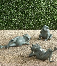 three metal frog statues sitting on top of a gravel covered ground next to a hedge