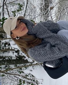 a woman in grey sweater and hat standing on snow covered ground with trees behind her