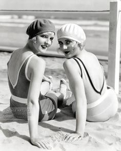 two women in bathing suits sitting on the sand at the beach, one wearing a hat