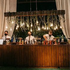 three men sitting at a bar with lights hanging from the ceiling and plants growing over them