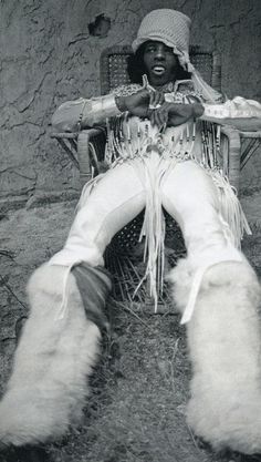 a man sitting in a chair with his legs crossed and wearing white furs on the ground