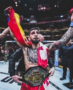 a man standing in a ring holding up a flag and a wrestling belt with his hands on his hips