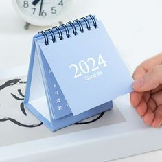 a hand is holding a blue calendar on a desk next to an alarm clock,