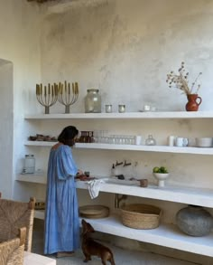 a woman in a blue dress standing next to a shelf filled with vases and bowls