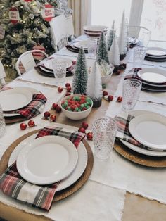 a table set for christmas with plates, silverware and small trees in the background