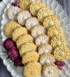 a white plate topped with lots of cookies next to purple and red flowers on top of a wooden table