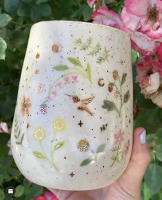 a hand holding up a white glass with flowers and butterflies painted on the outside, in front of some pink roses