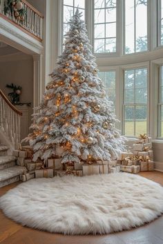 a white christmas tree with lights and presents in front of a large window on a wooden floor