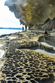 two people are standing on the edge of a rock formation near water and sand with holes in it