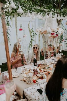 two women sitting at a table with food and drinks in front of them, surrounded by greenery