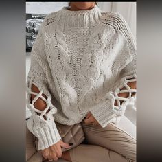 a woman wearing a white sweater with cutouts on the sleeves and cuffs, sitting in front of a window