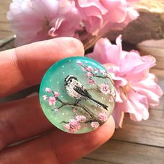 a hand holding a glass caboch with a bird on it and pink flowers in the background