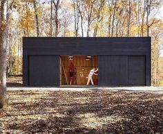 two people are walking out of a black building in the woods with autumn leaves on the ground