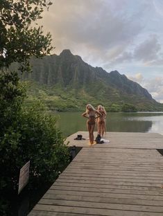 two women in bathing suits are walking on a dock near the water with mountains in the background