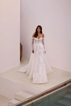 a woman in a white wedding dress standing next to a pool wearing an off the shoulder gown